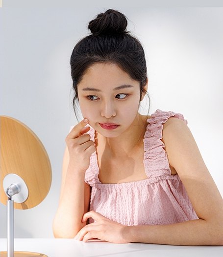 분홍 프릴 원피스를 입은 여성이 책상에 앉아 손가락으로 뺨을 만지며 탁상 거울을 바라보는 모습