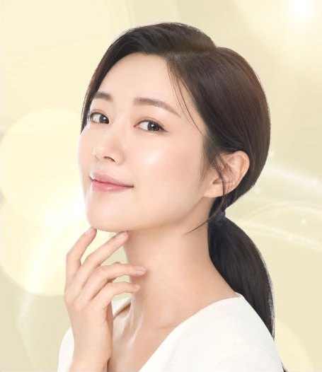 긴 머리를 하나로 묶은 여성 모델이 손끝을 턱선에 대고 옅게 미소 짓는 상반신 컷