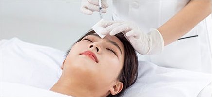 흰 시술대에 누운 여성의 이마에 라텍스 장갑을 낀 시술자가 주사기로 스킨부스터를 주입하는 모습