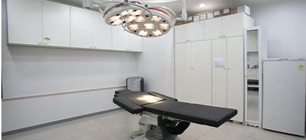무영등이 달린 시술실 내부 — 검은 시술 베드와 흰 수납장, 소형 냉장고와 거울장이 놓인 모습